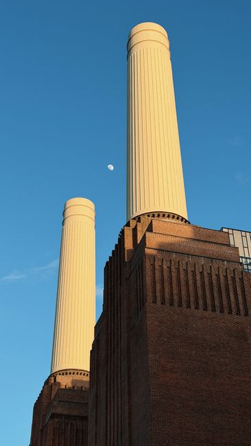 A close-up view of the top section of Battersea Power Station with two large, white, cylindrical chimney stacks set against a clear blue sky. The building's brick facade is visible at the base, featur