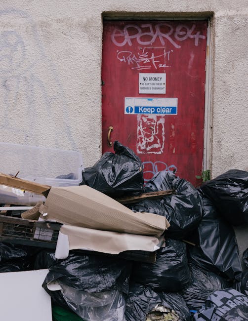 A pile of discarded household rubbish and debris arranged against an outdoor brick wall, including broken wooden furniture such as chairs and slats, a damaged microwave or small appliance with its doo