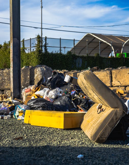 A pile of mixed household waste and debris, including black plastic rubbish bags, a yellow plastic storage container, and part of an old, worn car tire, all situated on a gravel surface next to a ston
